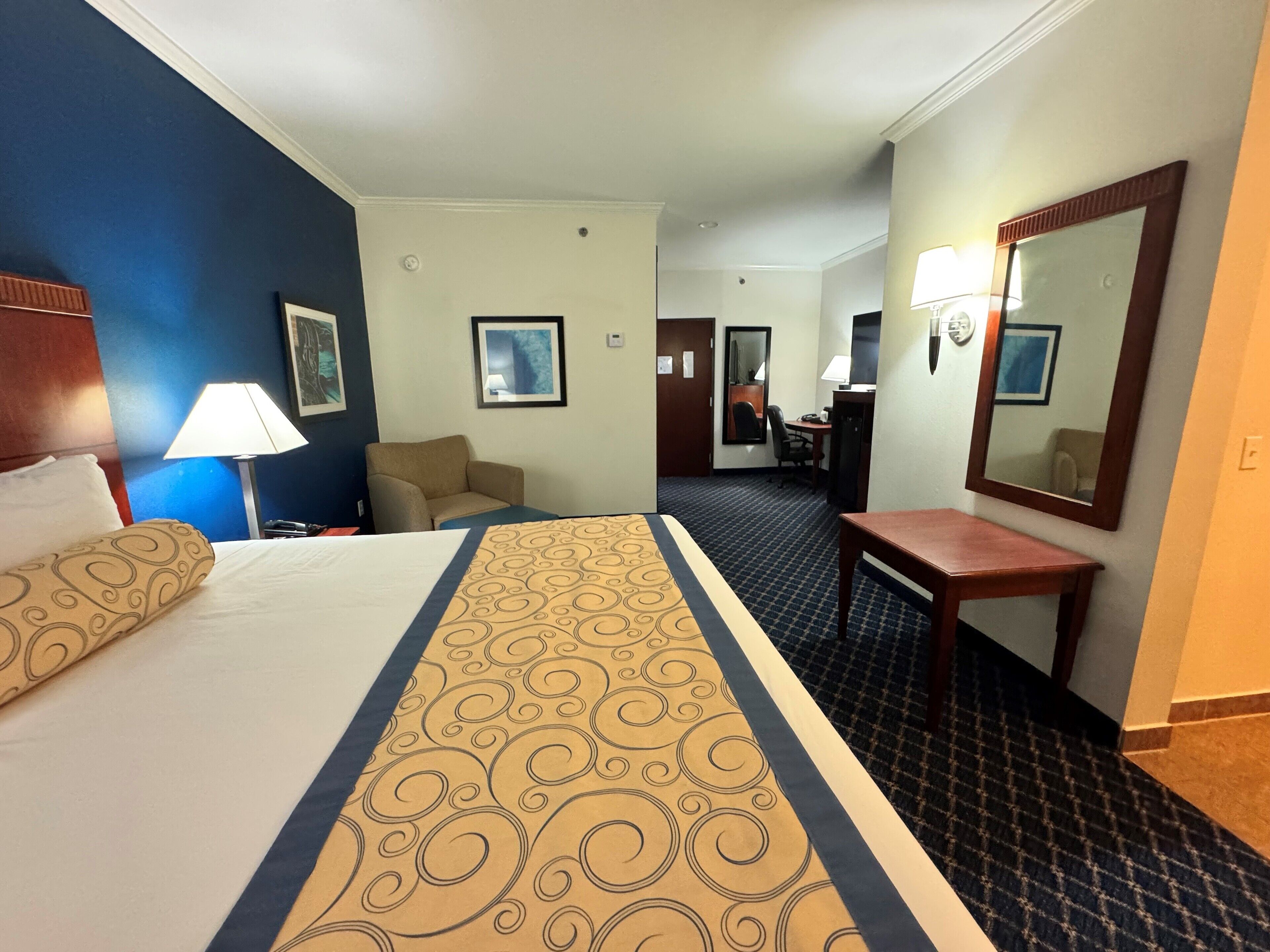 Habitación de hotel con cama, escritorio, silla, espejo y lámpara.