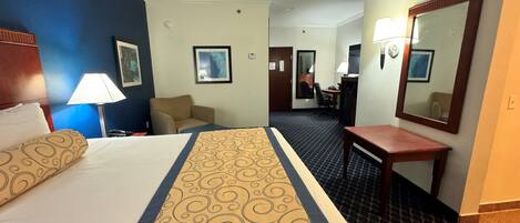 A hotel room with a bed, a desk, a chair, a mirror, and a lamp.