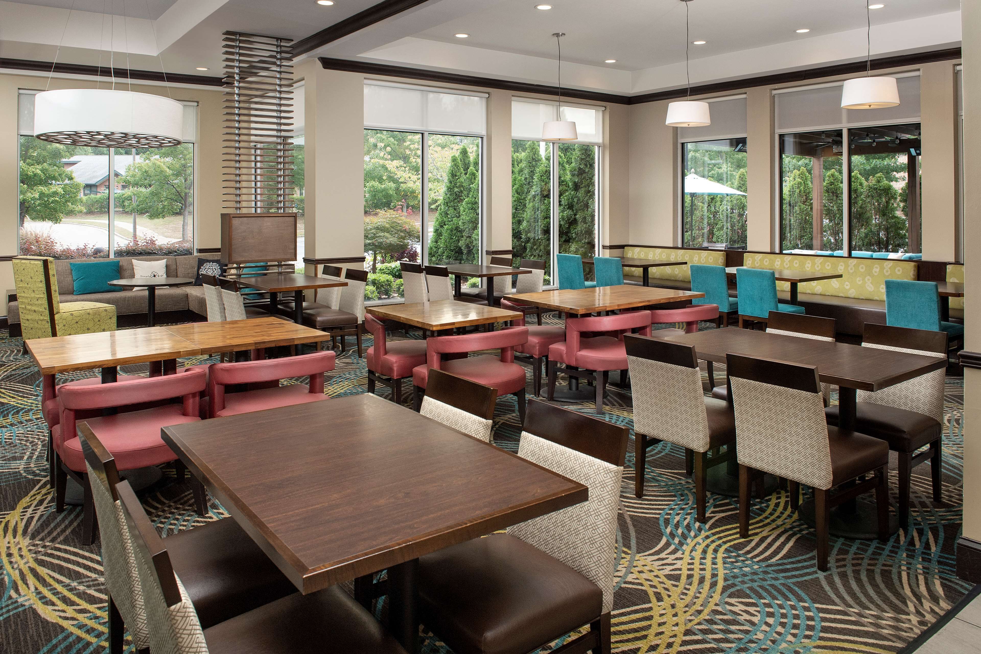 A hotel dining area with multiple tables and chairs, large windows with a view of greenery, and a patterned carpet.