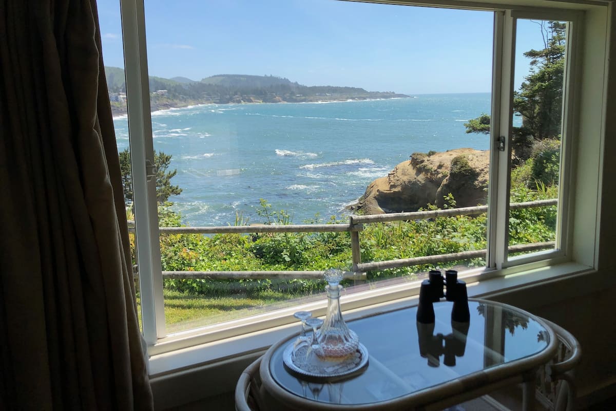 A room with a large window offering a view of the ocean and cliffs.