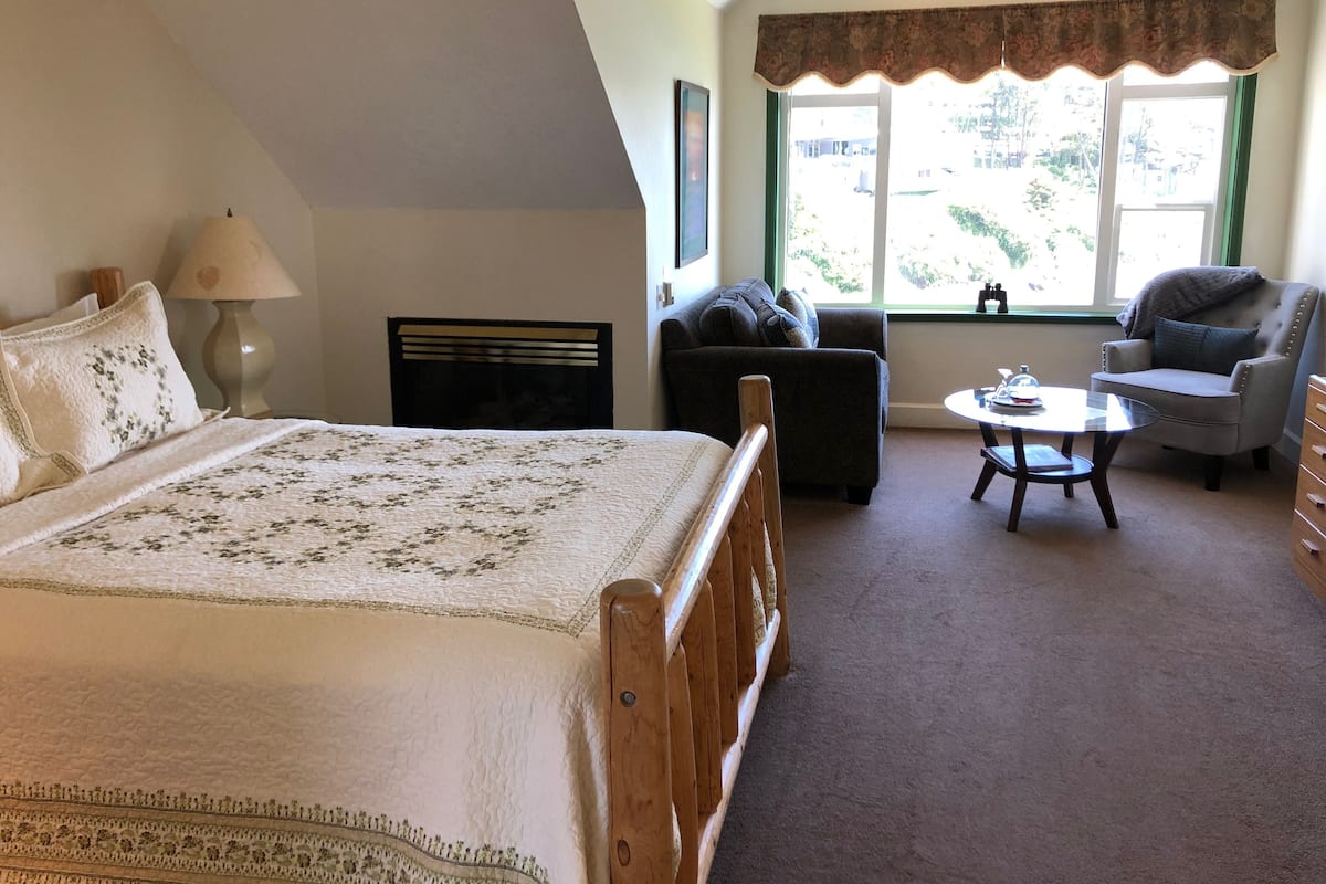 A bedroom with a large bed, a chair, a small table, and a window with a view of greenery.
