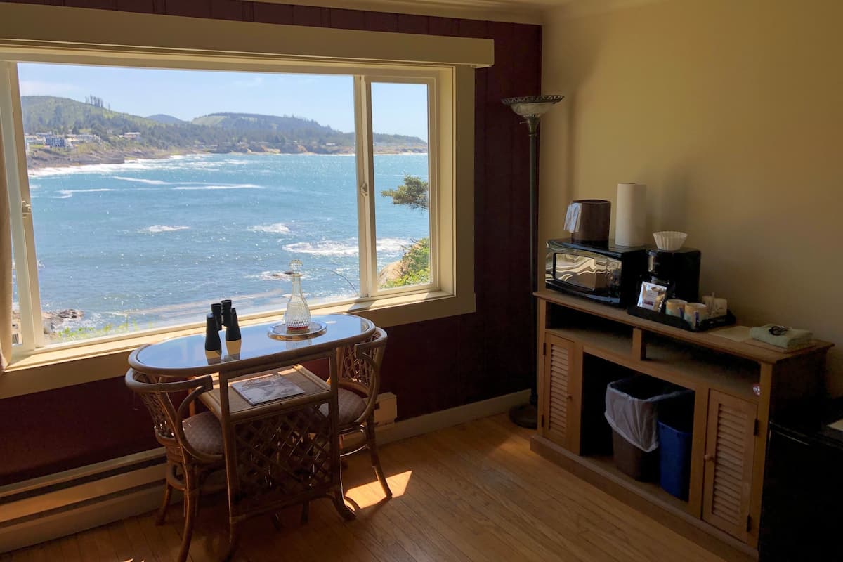 A room with a large window overlooking the ocean, a small table with chairs, and a coffee station.