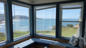 Habitación de esquina con ventanal panorámico que da a la costa, un banco de madera y una mesa con un vaso.