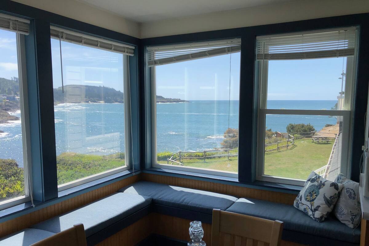 A corner room with a bay window overlooking a coastal view, a wooden bench, and a table with a glass.