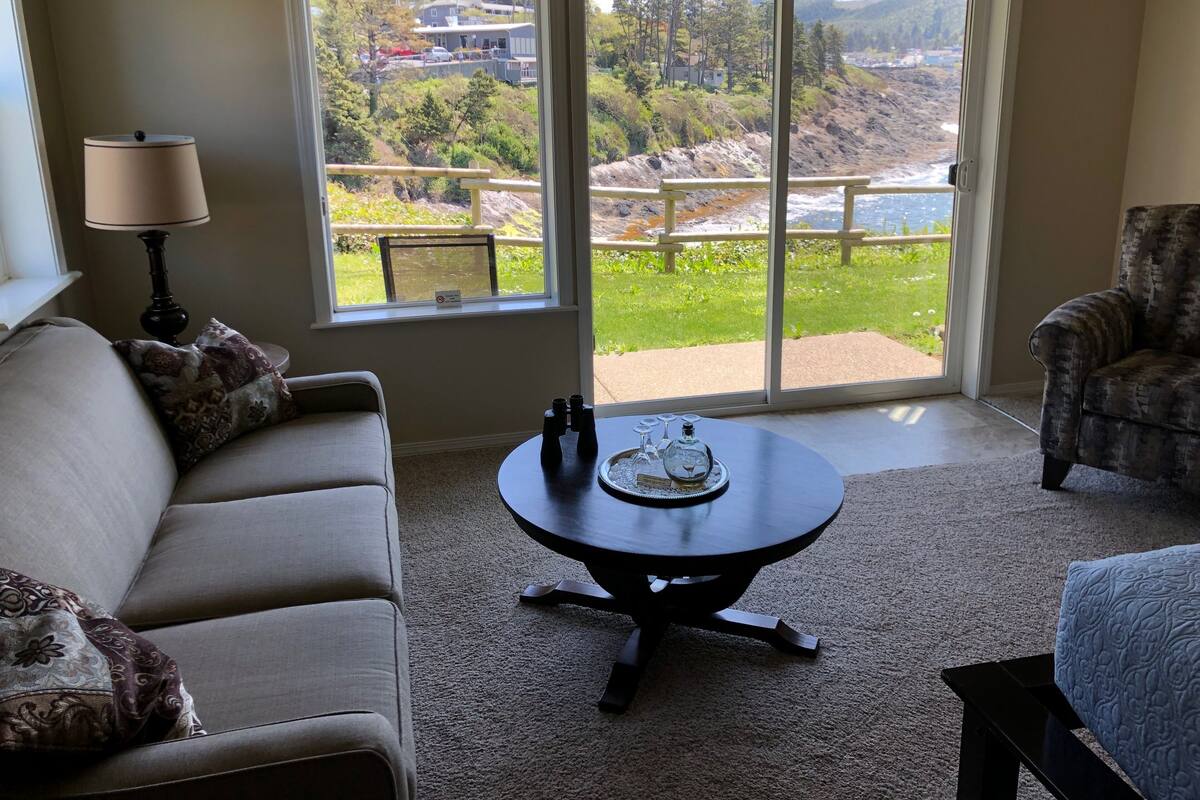A living room with a sofa, armchair, and a round table with a tray of glasses and a bottle. A large window offers a view of a green landscape and a body of water.