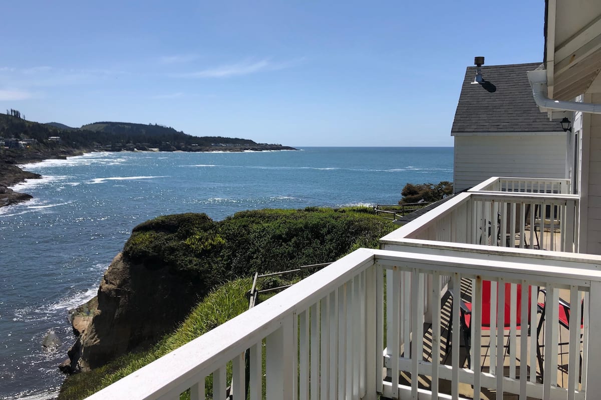 A coastal view from a balcony with a white railing, overlooking the ocean and a rocky shoreline.