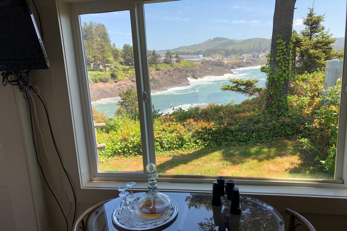 A table with a glass top, a decorative glass piece, and binoculars, positioned by a window with a coastal view.