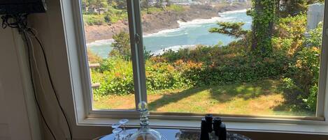 A table with a glass top, a decorative glass piece, and binoculars, positioned by a window with a coastal view.