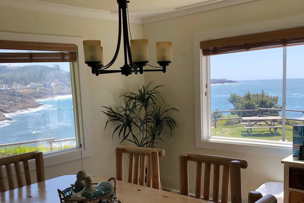 A dining area with a wooden table, chairs, a chandelier, and large windows offering ocean views.