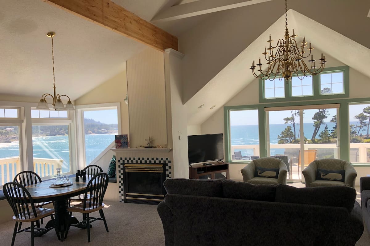 A spacious living room with a fireplace, a dining area with a table and chairs, and large windows offering ocean views.