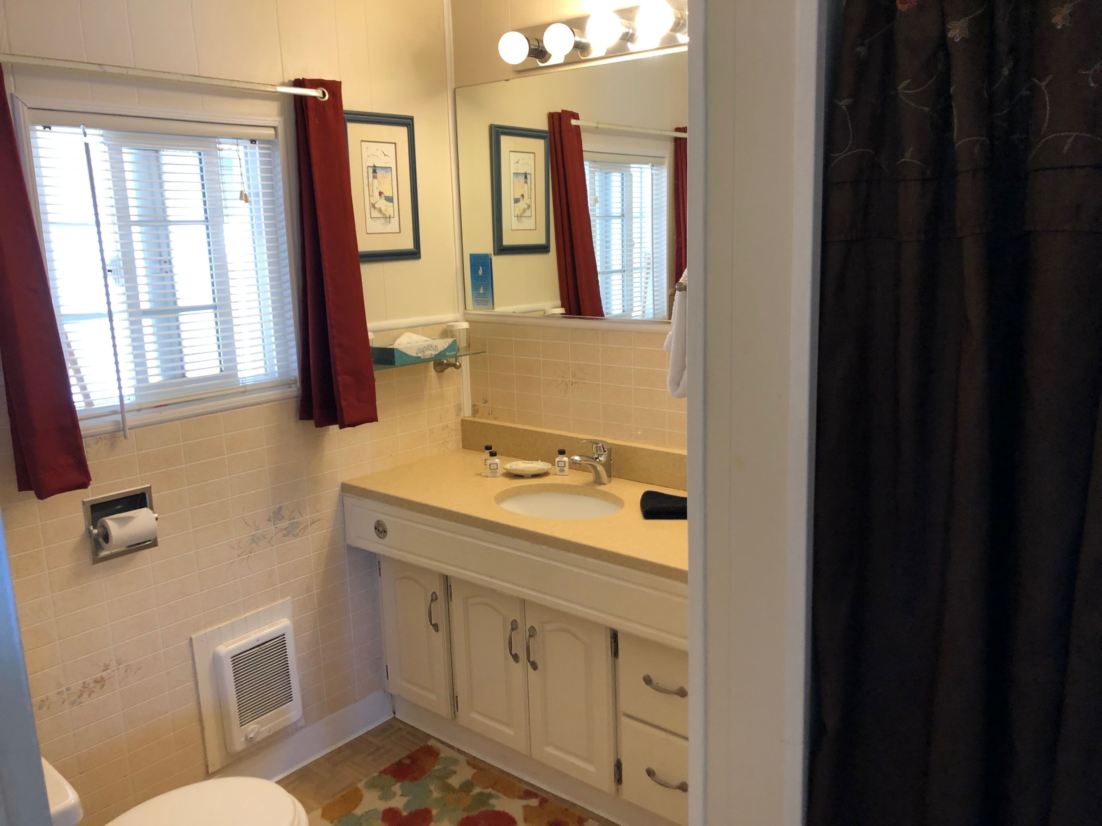 A bathroom with a toilet, sink, and shower area. There are curtains, a mirror, and a window with blinds.