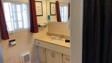 A bathroom with a toilet, sink, and shower area. There are curtains, a mirror, and a window with blinds.