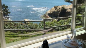 Habitación con un amplio ventanal que da a la costa, una mesa redonda de cristal con una botella y una copa, y una silla.