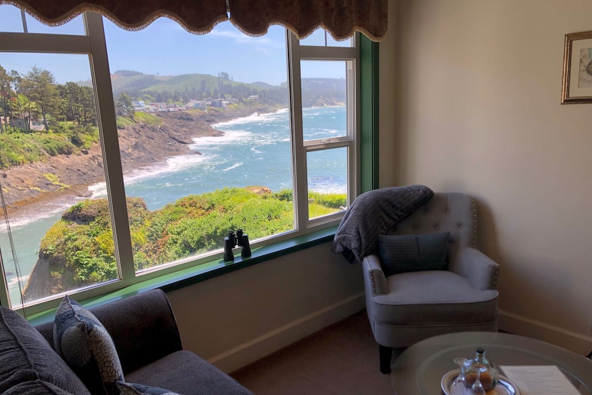A room with a large window overlooking a coastal landscape, a comfortable armchair, a sofa, and a small table with a decorative item.