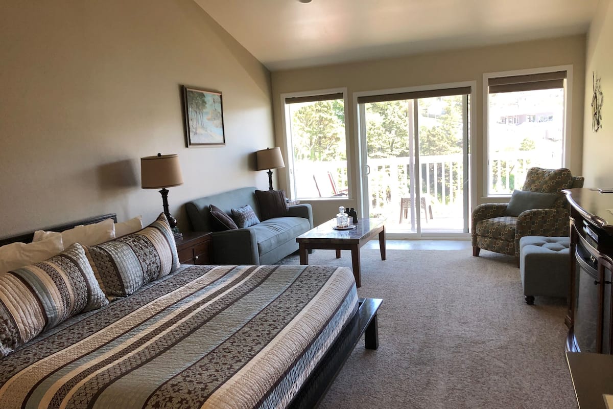 A spacious living room with a large bed, a sofa, a chair, and a table. There is a sliding glass door leading to a balcony.
