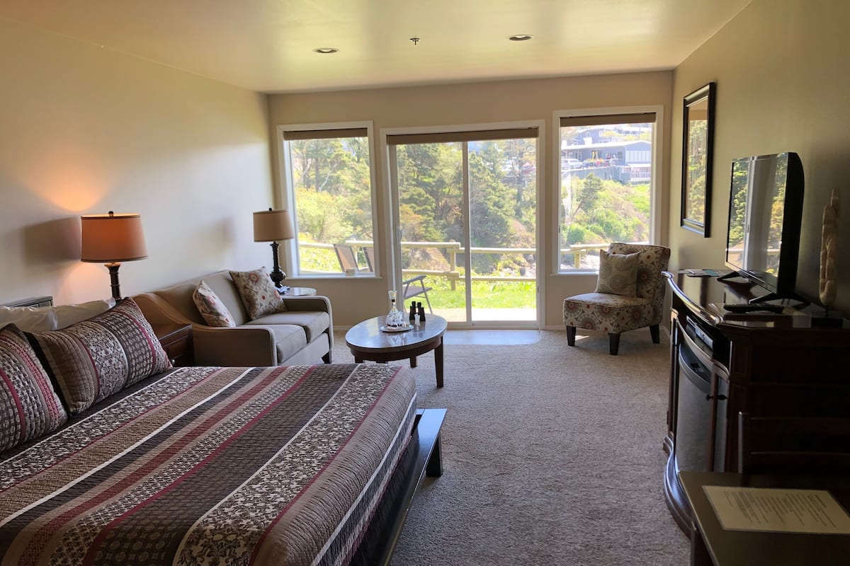 A spacious bedroom with a large bed, a sofa, a chair, a small table, and a dresser. There are large windows with a view of greenery outside.