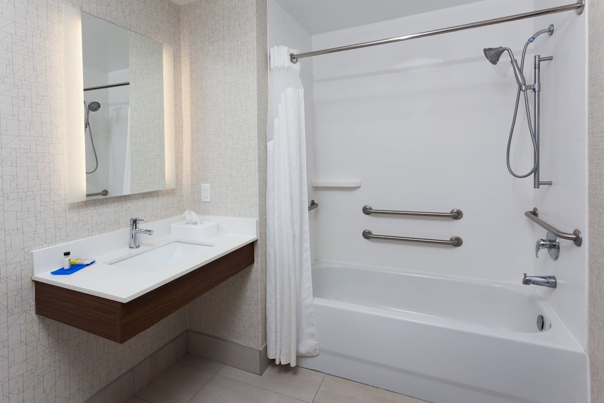 Un baño moderno con lavamanos, espejo y bañera blancos. Incluye un cabezal de ducha, barras de apoyo y un perchero.