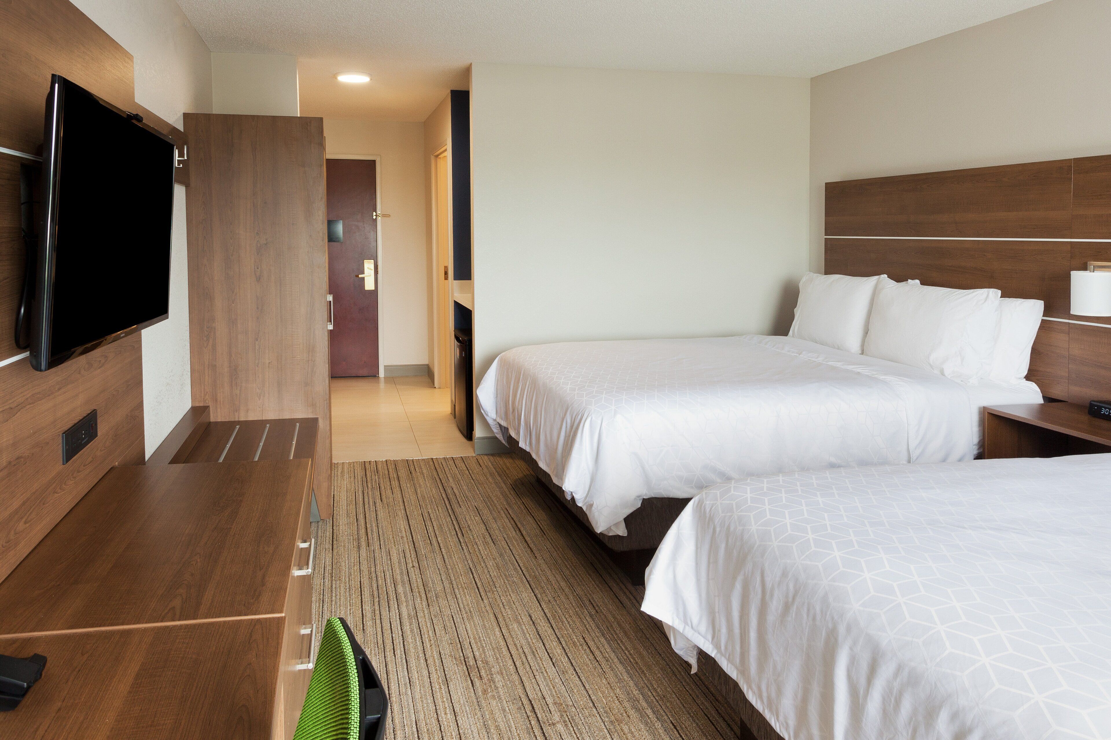 A hotel room with two beds, a flat-screen TV mounted on the wall, a wooden desk, and a green chair.
