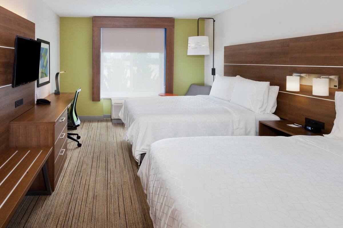 Habitación de hotel con dos camas, televisión, escritorio con silla, lámpara y ventana con persianas.