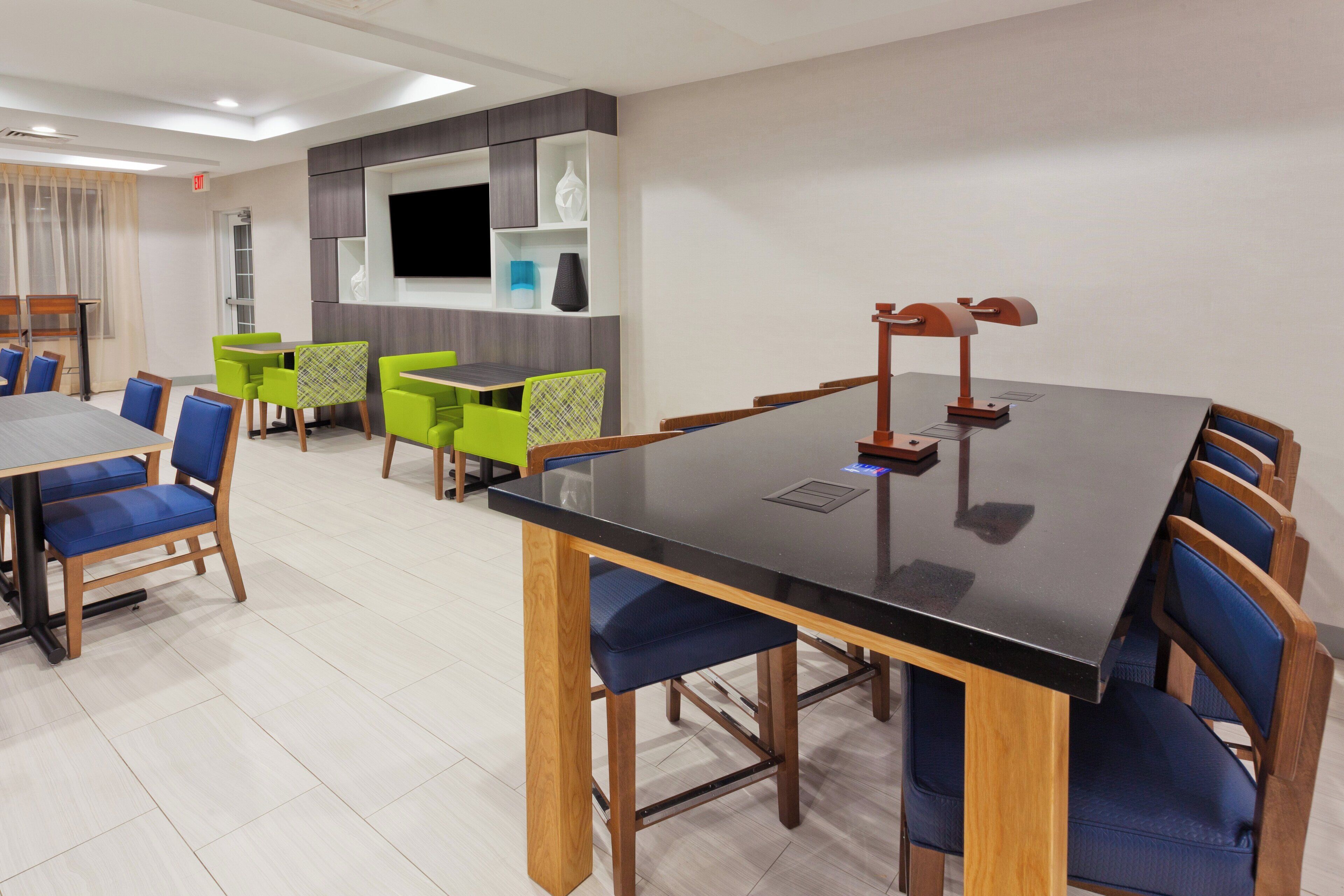 A hotel dining area with a large black table, blue and green chairs, and a television on the wall.