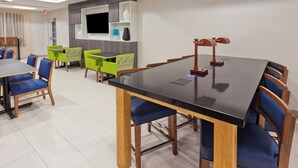 A hotel dining area with a large black table, blue and green chairs, and a television on the wall.