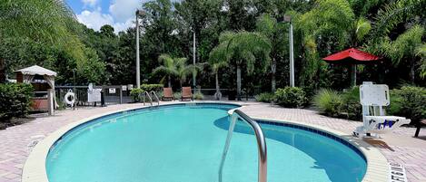 A circular pool with a metal ladder, surrounded by a brick walkway and lush greenery.