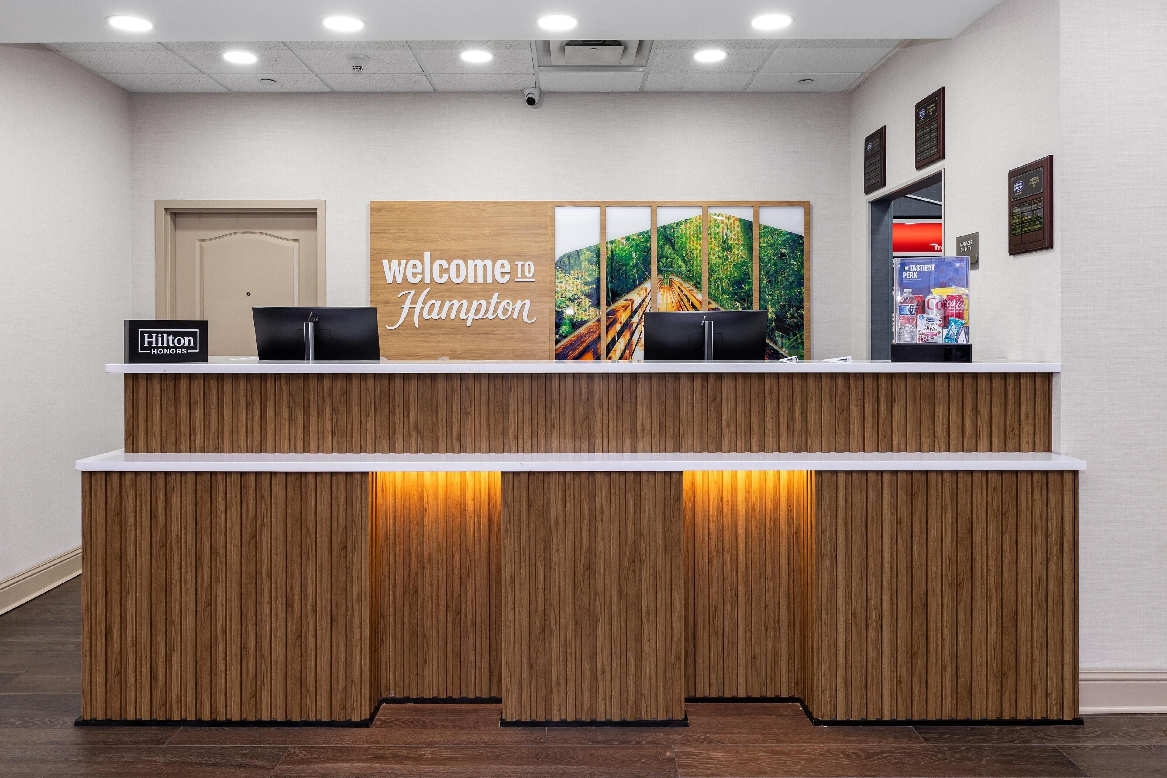 A reception desk with a 'Welcome to Hampton' sign, a 'Hilton Honors' sign, and a refrigerator stocked with beverages.