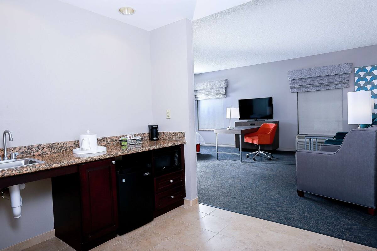 A compact hotel room with a kitchenette, a small dining area, a TV, and a seating area with a sofa and chair.