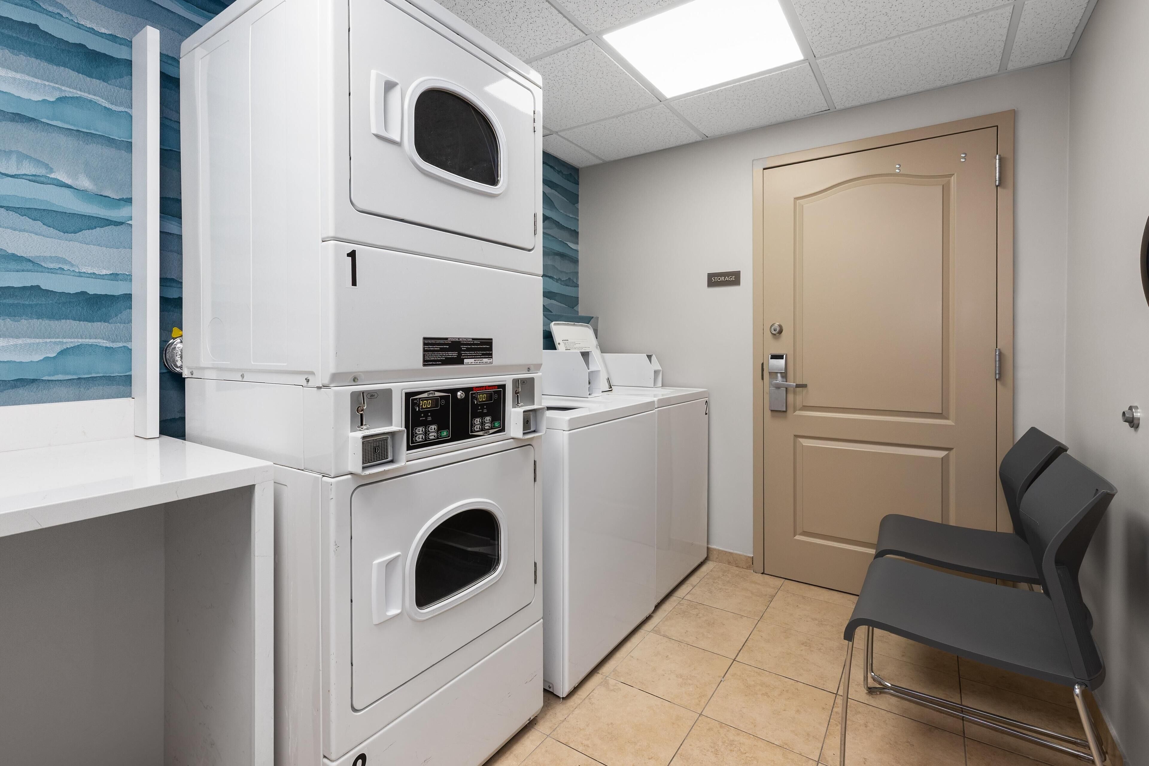 A laundry room with stacked washers and dryers, a storage sign, a bench, and a door.