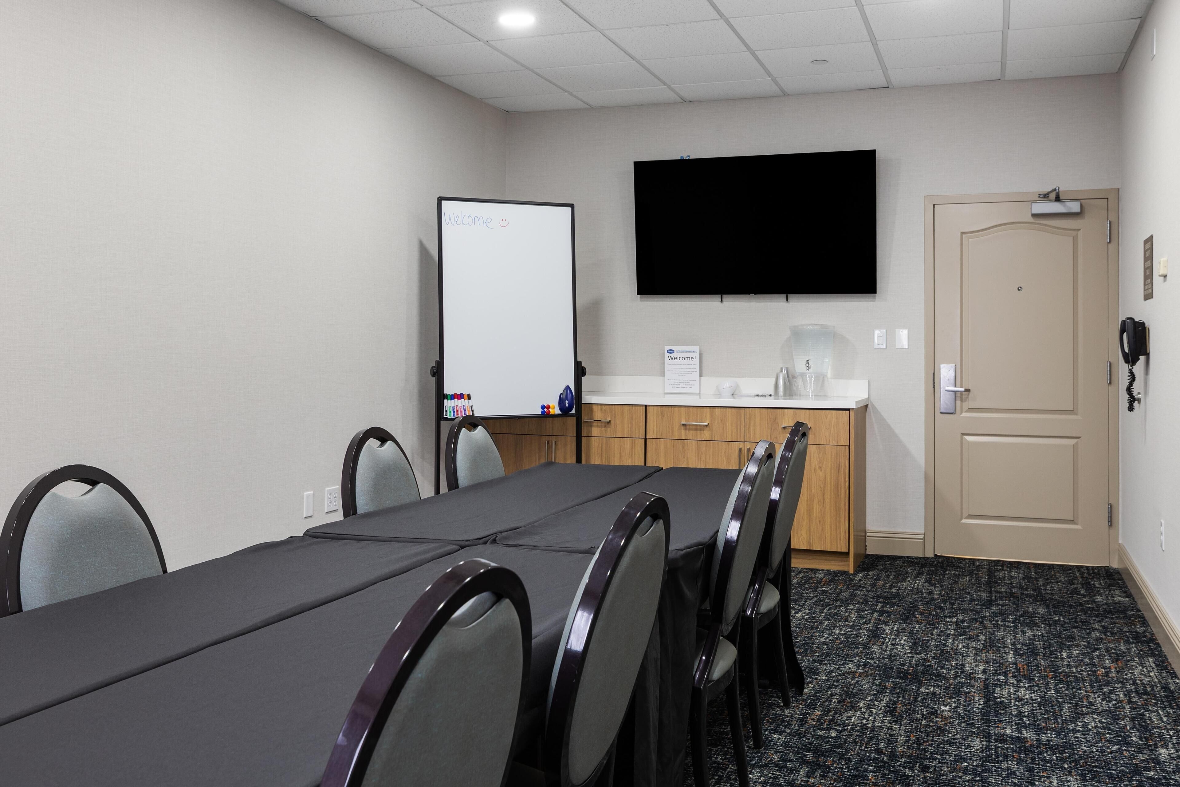 Un salón de conferencias con una larga mesa, sillas, una pizarra, un televisor de pantalla plana, una estación de café y una puerta.