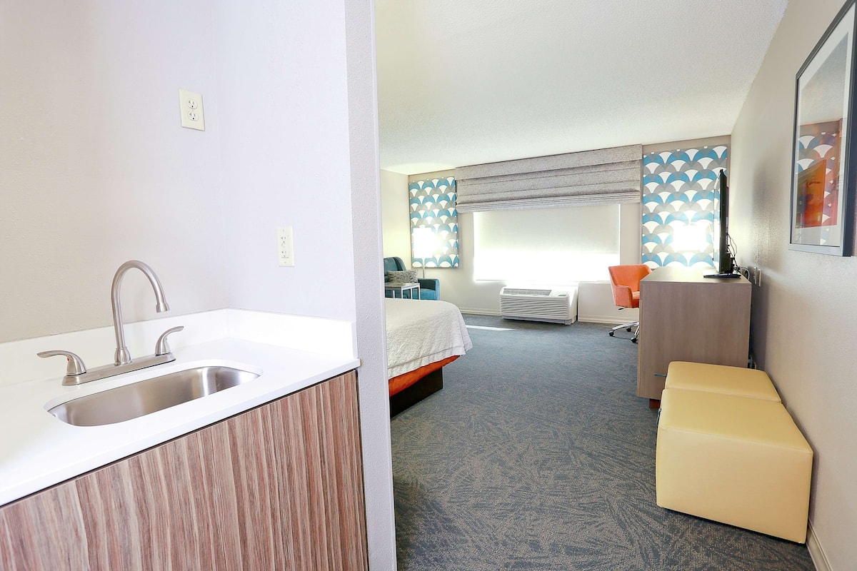 A hotel room with a modern bathroom, a bed, a desk, and a small seating area.