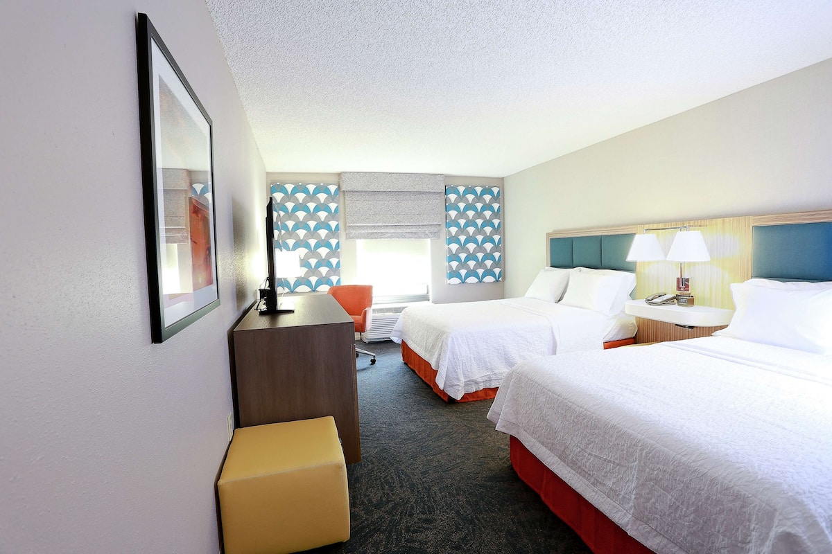 A hotel room with two beds, a desk, a chair, a small table with a lamp, and a window with blinds.