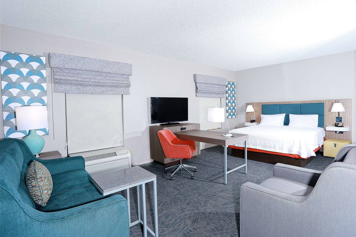 A hotel room with a bed, desk, two chairs, a TV, and a window with blinds.