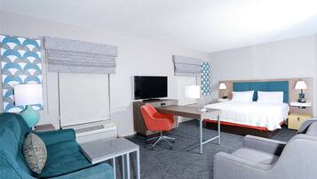 A hotel room with a bed, desk, two chairs, a TV, and a window with blinds.