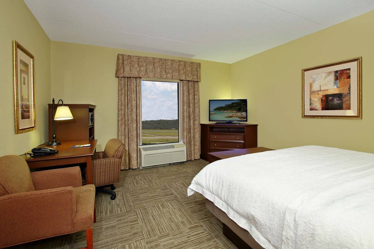 A hotel room with a bed, a desk with a lamp, a chair, a television, and a window with curtains.