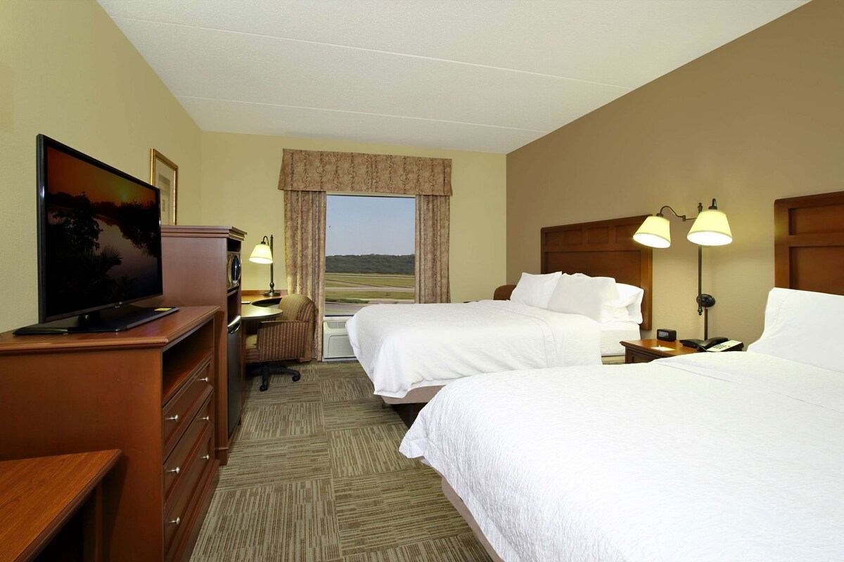 Ein Hotelzimmer mit zwei Betten, einem Schreibtisch mit Fernseher, einer Lampe und Blick auf eine Landschaft.