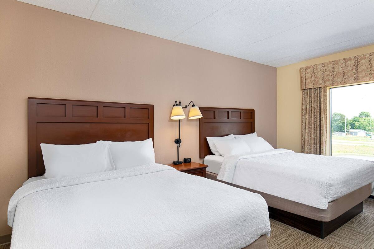 A hotel room with two beds, a nightstand with a lamp, and a large window with curtains.