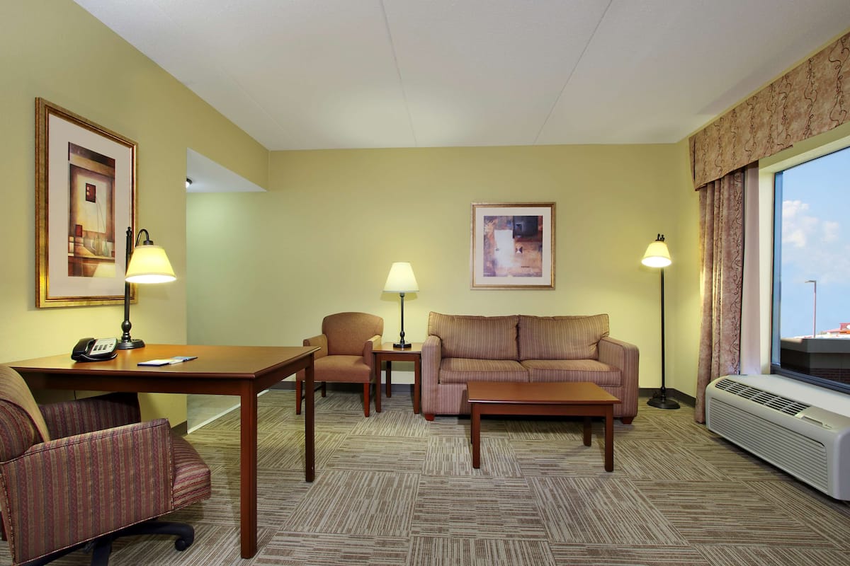 A hotel room with a sofa, armchair, small table, and a desk with a lamp.