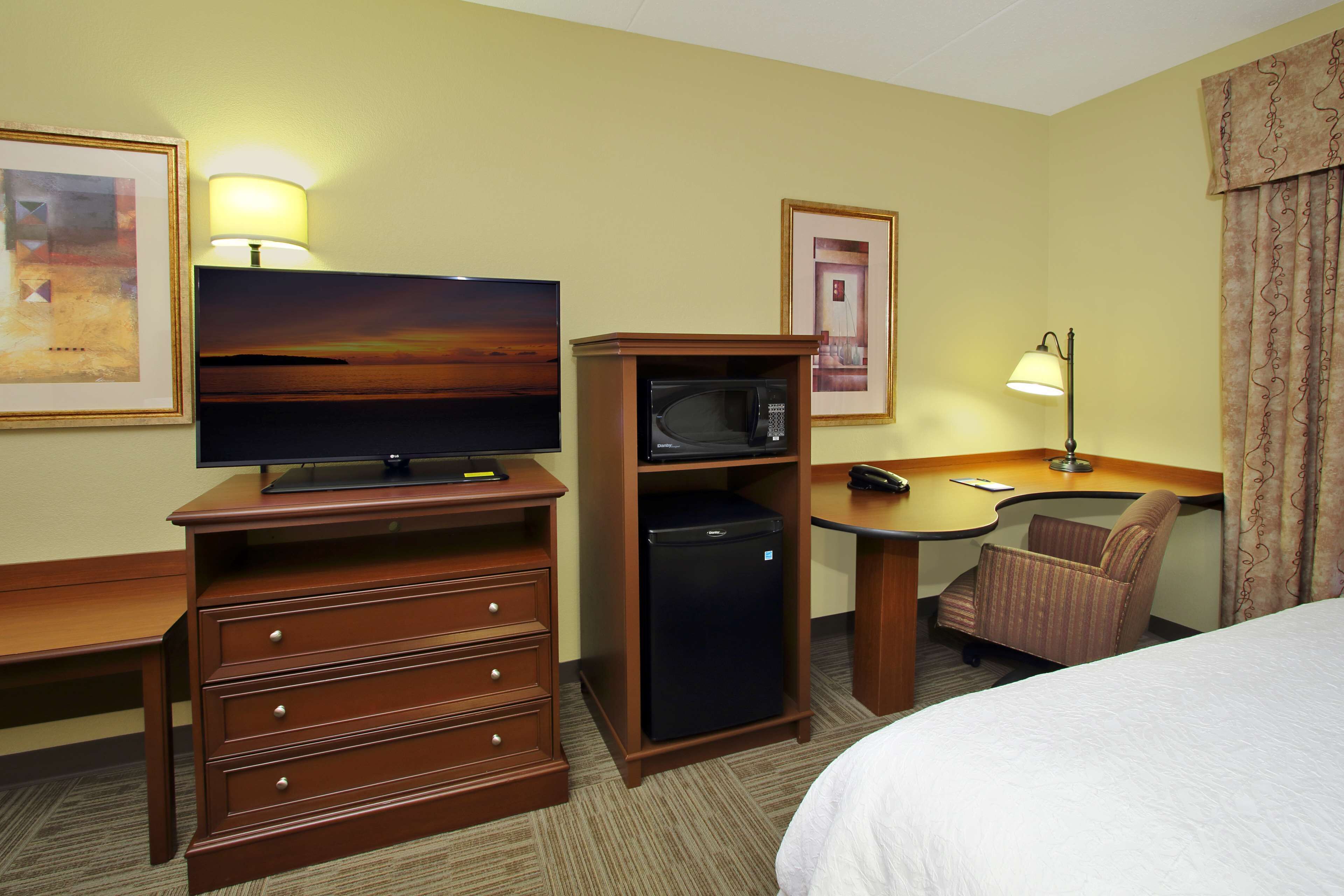 Una habitación de hotel con una cama, un escritorio con lámpara, un televisor, un refrigerador y una silla.