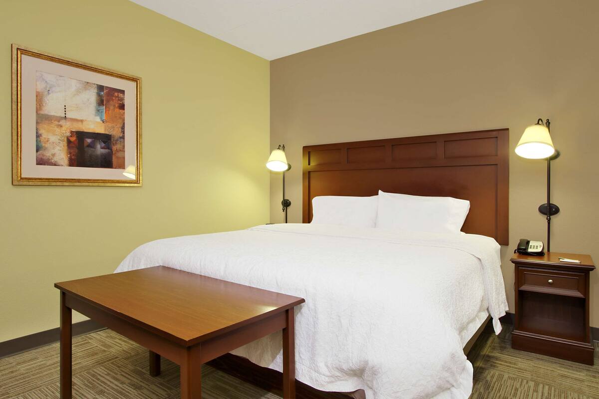A hotel room with a large bed, bedside lamps, a small table, and a framed picture on the wall.