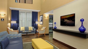 A hotel room with a sofa, armchairs, a yellow ottoman, a TV, and decorative vases. - Hampton Inn & Suites Madisonville (Madisonville)