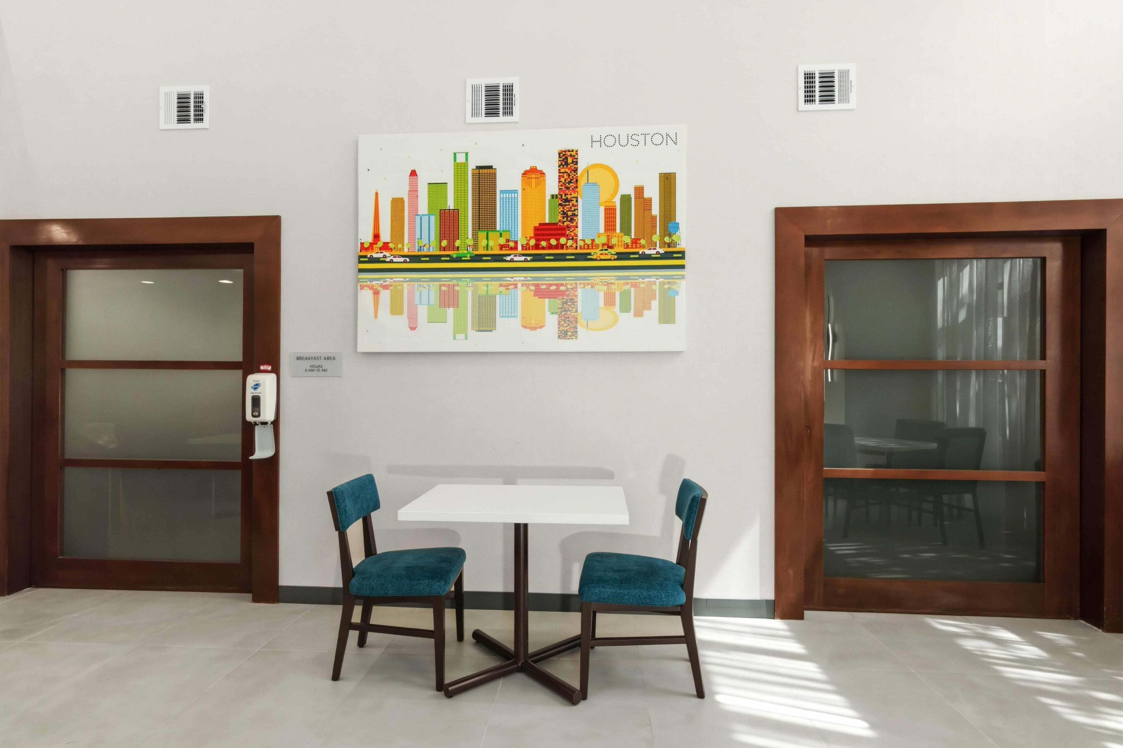 A hotel room with a white table and two teal chairs, a cityscape artwork on the wall, and two doors with frosted glass panels.