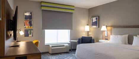 A hotel room with a large bed, a desk with a computer, a chair, a TV, and a window with a view.