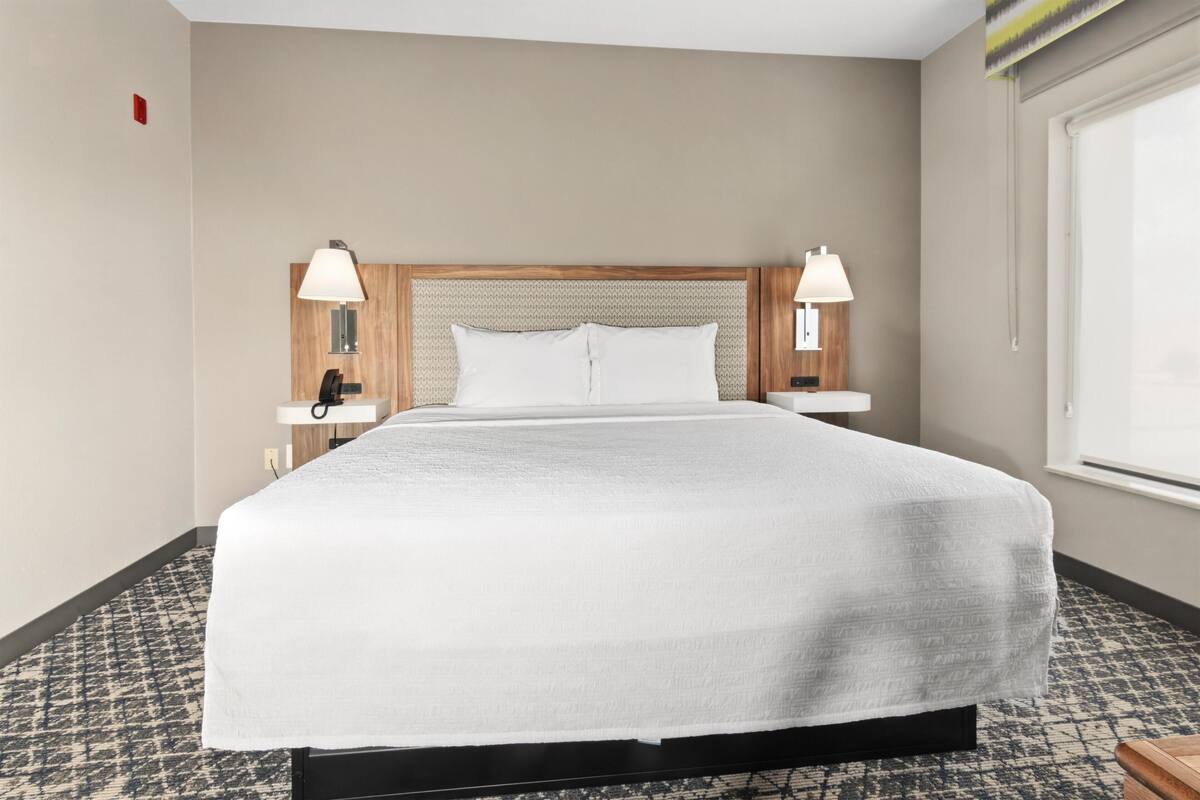 A hotel room with a large bed, two bedside tables with lamps, a telephone, and a window with blinds.