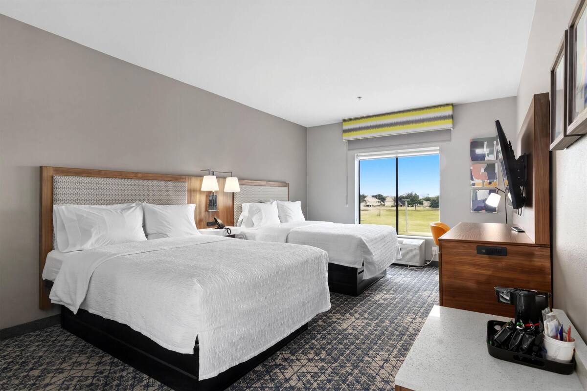 A hotel room with two beds, a desk with a coffee maker, a TV, and a large window with a view of the outdoors.