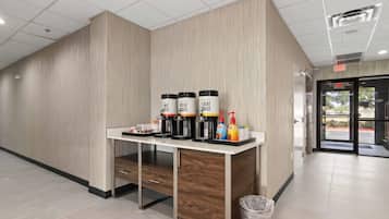 Una estación de café con tres dispensadores, un cesto de basura y una puerta que da a un espacio al aire libre.