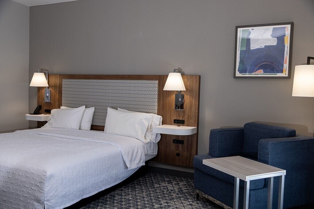A hotel room with a large bed, two bedside tables with lamps, a small desk, and a blue armchair.