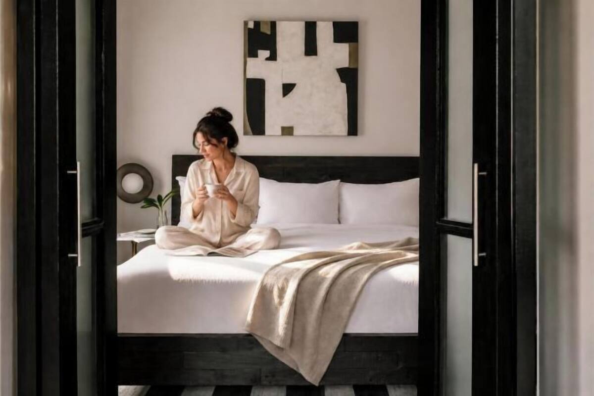 Une personne est assise sur un lit dans une pièce au design moderne, avec une table de chevet sur laquelle se trouve une lampe, et une œuvre d’art accrochée au mur.