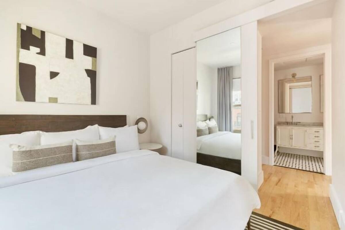Une chambre d’hôtel comprenant un lit, une table de chevet, un miroir, une salle de bain avec un lavabo et un miroir, et un plancher en bois.