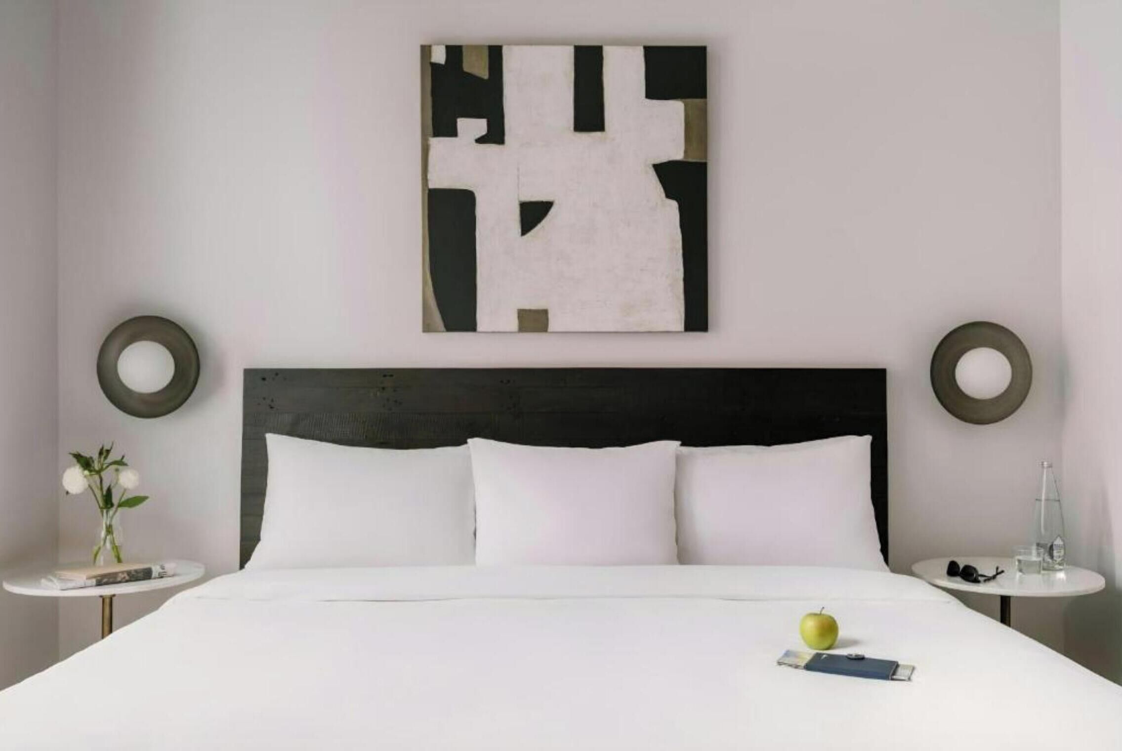 A hotel room with a large bed, two bedside tables, a wall-mounted abstract artwork, and a small round table with a vase of flowers.